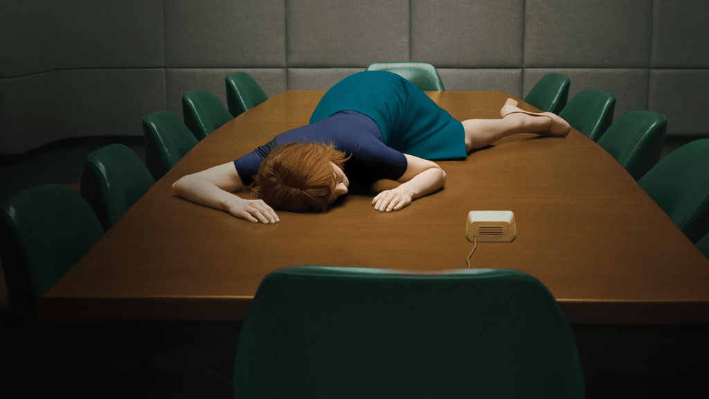 A person with red hair dressed in a blue outfit is lying face down on a wooden conference table, surrounded by green chairs.